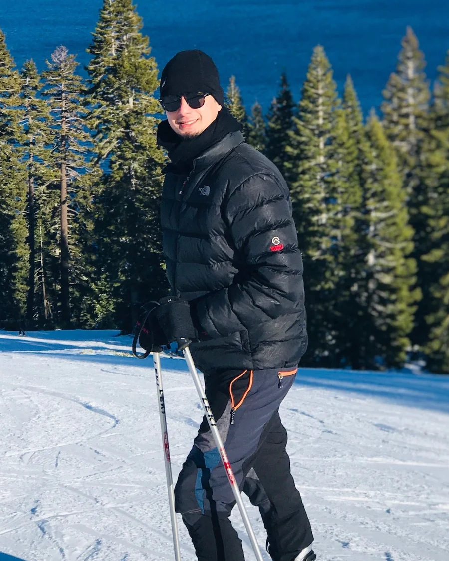 Avinash Koirala on the slopes above Lake Tahoe, in a black jacket and beanie with skis and poles.